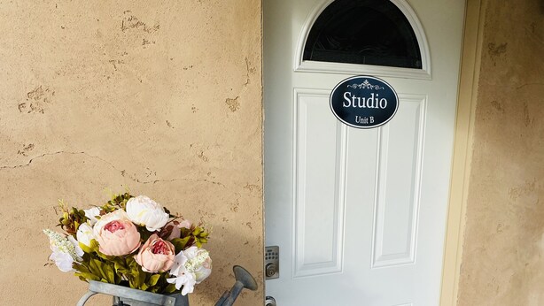 Un barril de madera con un farol y flores, una puerta con un cartel que dice "Studio Unit B", y un felpudo.