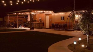 Un patio al aire libre con luces colgantes, mesas y sillas de madera, ideal para una comida relajada o encuentros sociales.
