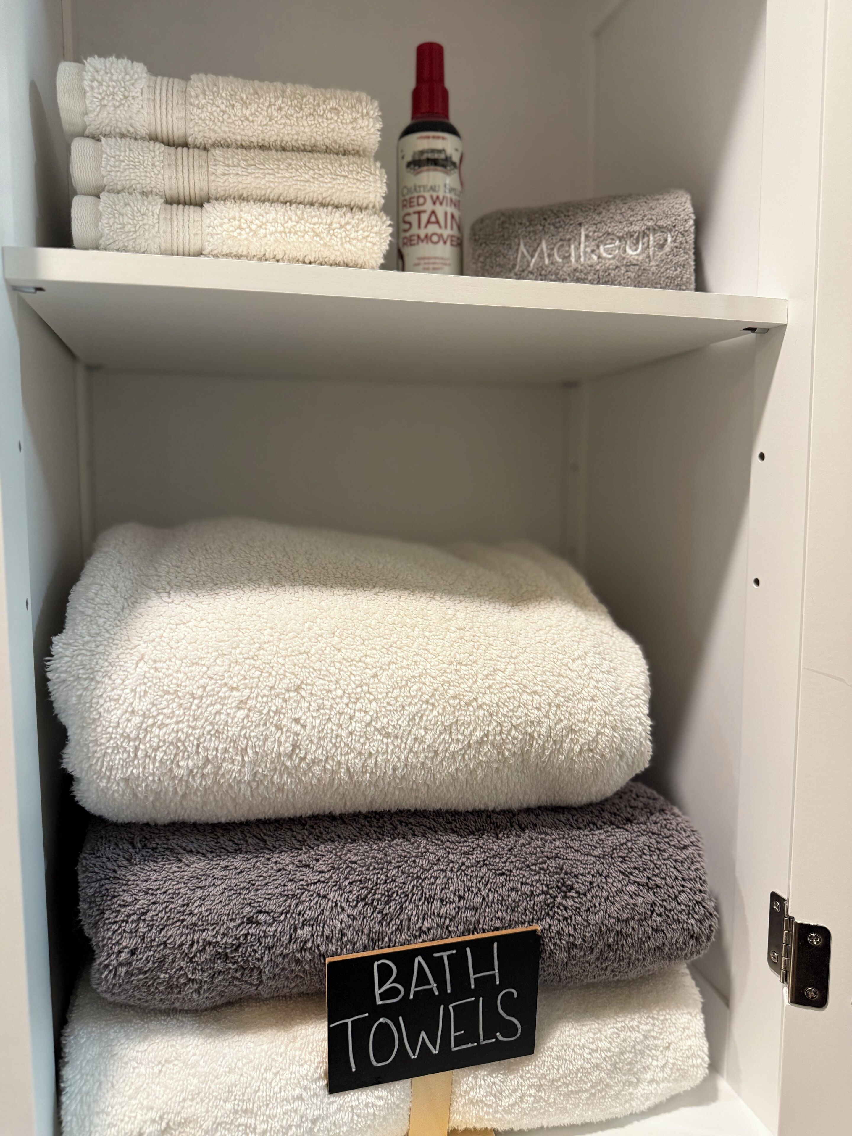Un espacio de almacenamiento con toallas dobladas ordenadamente en blanco y gris, un quitamanchas para vino tinto, y un cartel que dice 'TOALLAS DE BAÑO'.