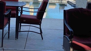 Zona de piscina con sillas de descanso y sombrillas, rodeada por una valla y palmeras.