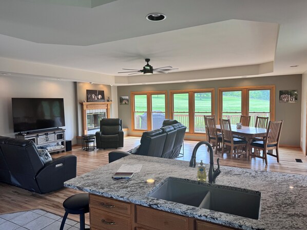 A spacious living area with a large TV, comfortable seating, a fireplace, and a view of a green landscape through large windows.