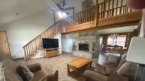 A spacious living room with a stone fireplace, wooden staircase, and a flat-screen TV.