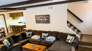 Ein gemütliches Wohnzimmer mit einem braunen Ecksofa, einem Holztisch und Kissen mit Muster. Im Hintergrund ist eine Treppe zu einer weiteren Ebene zu sehen.