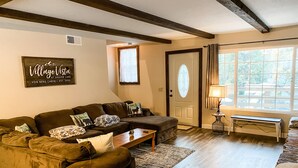 Ein gemütliches Wohnzimmer mit einer Eckcouch, einem Couchtisch und einer Lampe. Eine Tür mit Fenster und ein Schild an der Wand sind ebenfalls zu sehen.