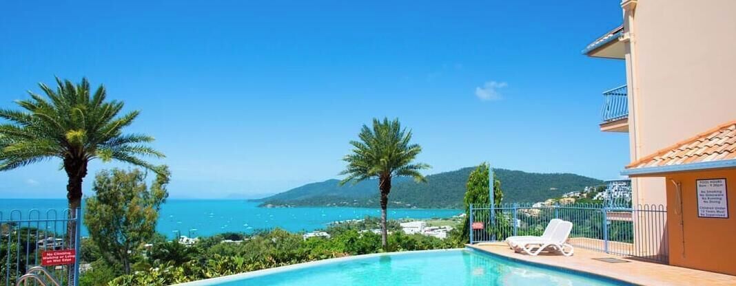 A pool area with a view of the sea and hills, palm trees, and a white lounge chair.