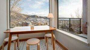 Ett rum med ett träbord och fyra pallar, en lampa och ett stort fönster med utsikt över en snötäckt stad.