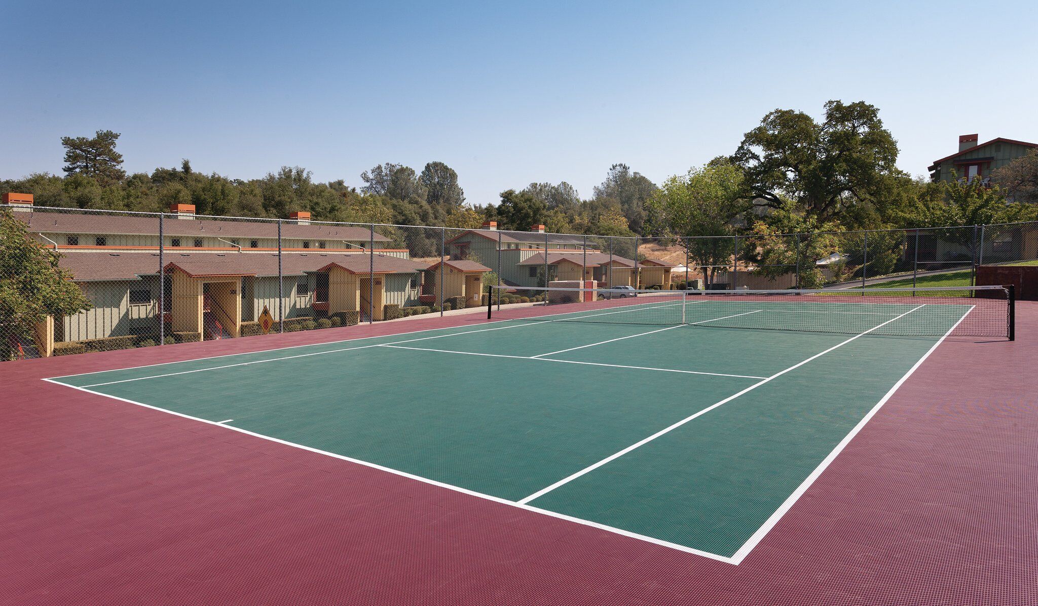 Un court de tennis avec un filet, entouré d’une clôture, et situé à côté d’une rangée de petits bâtiments.