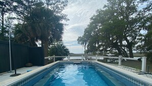 Piscina rectangular con muelle blanco y borde decorativo, rodeada de árboles y una valla de madera.
