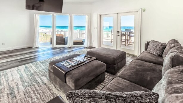 Ein geräumiges Wohnzimmer mit einem dunkelgrauen Sofa, einem Couchtisch und einem gemusterten Teppich. Zwei Schiebeglasfenster führen zu einem Balkon mit Blick auf den Strand.