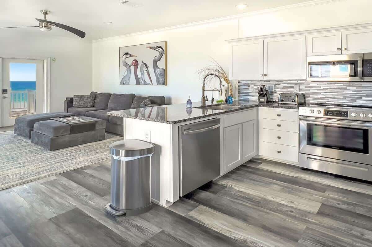 A modern kitchen with stainless steel appliances, a grey sofa, and a large window overlooking the ocean.