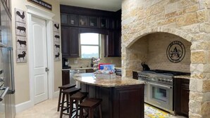 A kitchen with a stone fireplace, dark wooden cabinets, and a central island with stools. - NEW Hacienda Ranch W/ Resort Style Pool (Hempstead)