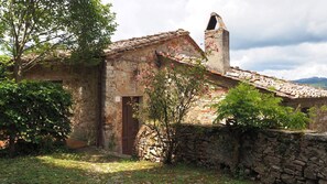 Un bâtiment rustique en pierre, doté d’un toit en tuiles, d’une cheminée et d’un petit jardin agrémenté d’un arbre et de plantes à fleurs.