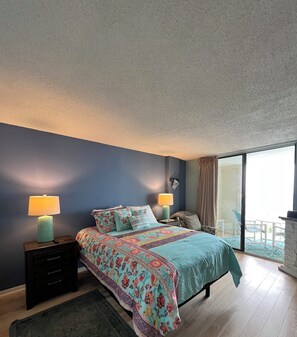A bedroom with a bed, two bedside tables, a nightstand, a window with curtains, and a view of the outdoors.