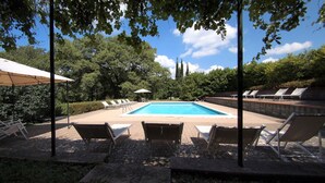 Piscina con sillas de descanso y sombrillas, rodeada de árboles y un muro de piedra.