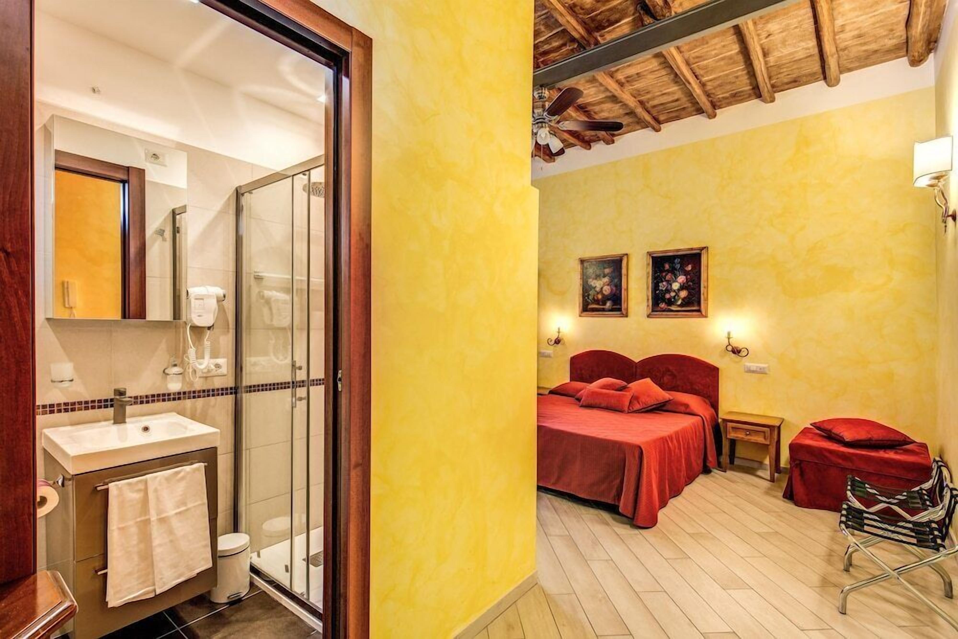 A bathroom with a glass shower enclosure, a sink, and a toilet. The bedroom has two beds with red bedding, a nightstand, and a chair.