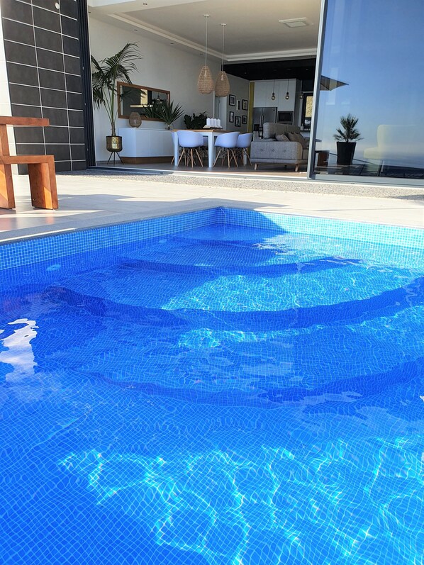 A modern indoor swimming pool with clear blue water, surrounded by a contemporary living area with a dining set and a sofa.