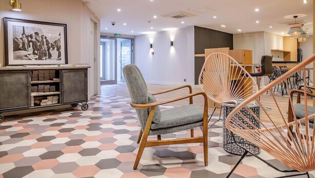 Un hall d’hôtel moderne avec un sol à motifs hexagonaux, une photographie en noir et blanc encadrée au mur, et un mobilier contemporain comprenant un fauteuil à revêtement gris et une chaise en rotin tressé.