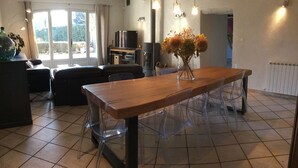 A dining area with a wooden table and clear chairs, a vase with dried flowers, and a television in the background.