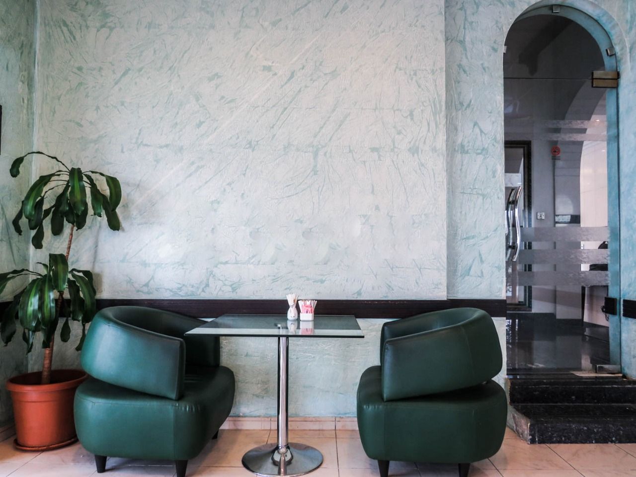 Eine Lobby mit Marmorwand, einem Glastisch, zwei grünen Stühlen, einem Blumentopf und einer Treppe im Hintergrund.