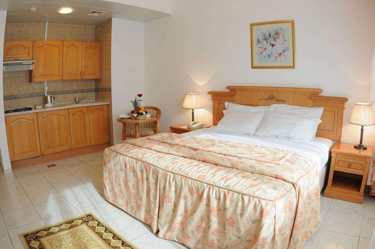 Een slaapkamer met een bed, nachtkastjes, een lamp, een schilderij en een keukenhoek met kastjes en een fornuis.