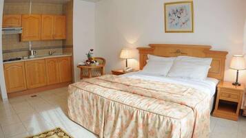 Une chambre à coucher comprenant un lit, des tables de chevet, une commode, une lampe, un tableau, et un coin cuisine avec des placards et une cuisinière.