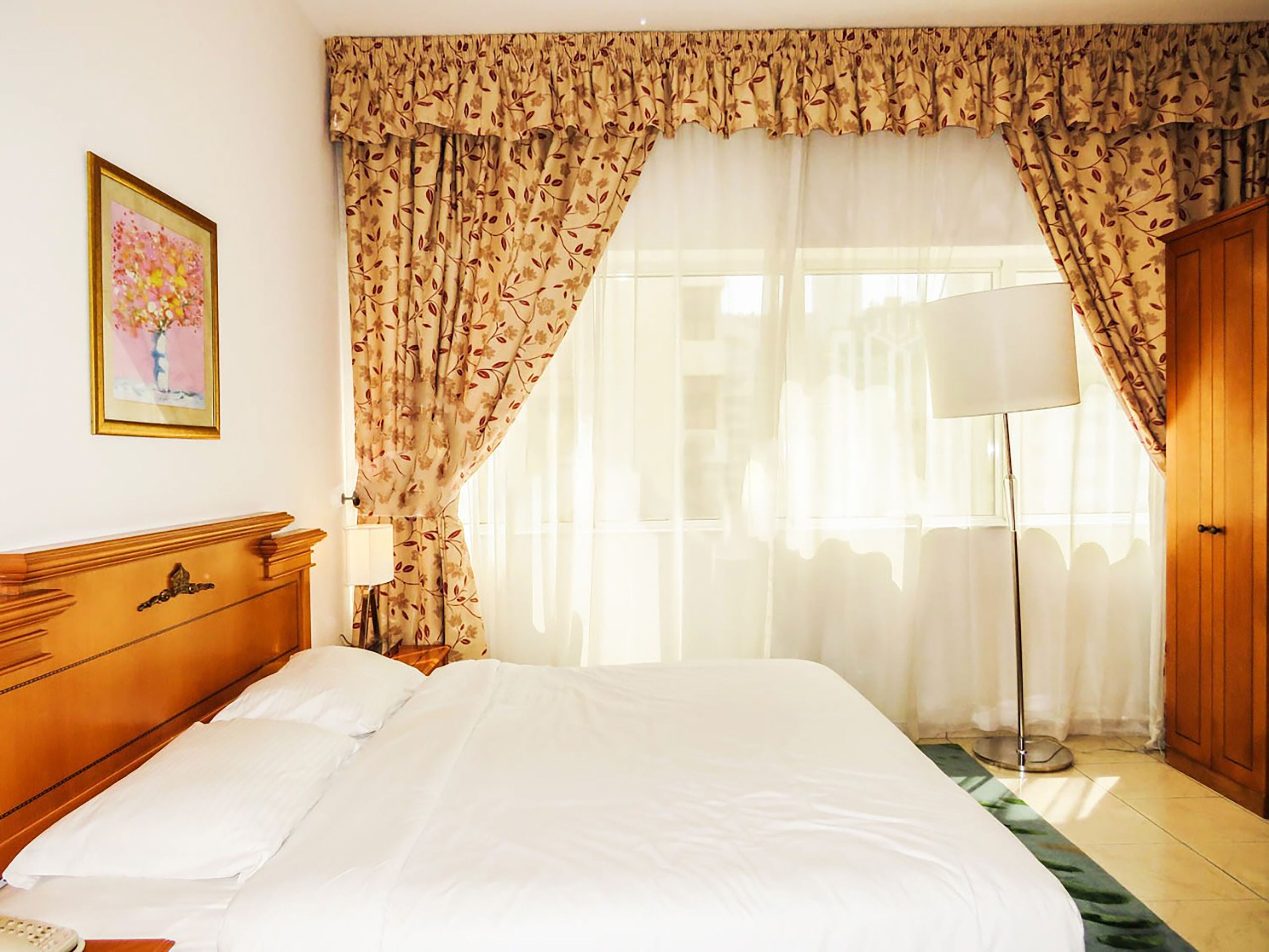 A hotel room with a large bed, a wooden headboard, a bedside table with a lamp, and floral curtains.