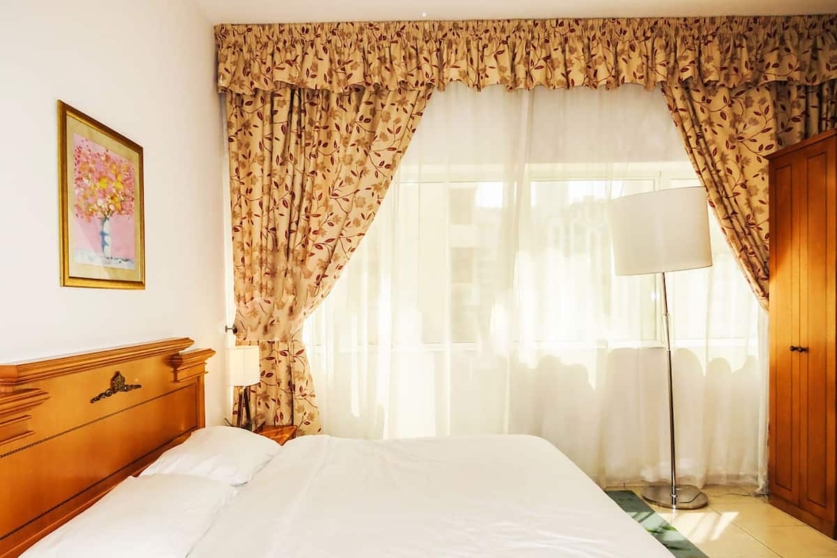 Une chambre d’hôtel avec un grand lit, une tête de lit en bois, une table de chevet avec une lampe et des rideaux à motifs floraux.