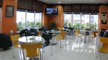 Ein Café mit runden Tischen und Stühlen, einem Fernseher an der Wand und großen Fenstern mit Jalousien.