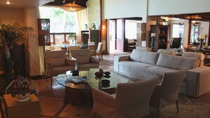 A spacious living area with a white sofa, armchairs, and a glass coffee table. There is a dining area in the background with a table and chairs.