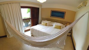 A bedroom with a bed, a hammock, a window with curtains, a painting on the wall, and an air conditioning unit.