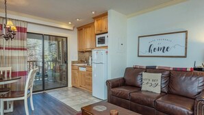 A living room with a brown leather sofa, a dining table with chairs, a kitchen area with wooden cabinets, a white refrigerator, and a sliding glass door leading to an outdoor area.