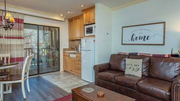 A living room with a brown leather sofa, a dining table with chairs, a kitchen area with wooden cabinets, a white refrigerator, and a sliding glass door leading to an outdoor area.