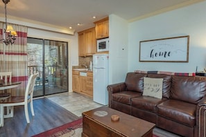 A living room with a brown leather sofa, a dining table with chairs, a kitchen area with wooden cabinets, a white refrigerator, and a sliding glass door leading to an outdoor area.