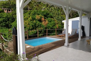 Ein überdachter Pool im Freien mit Holzterrasse, umgeben von üppiger Vegetation und einer weißen Pergola.