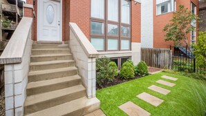A residential building with a brick facade, a white front door, a set of concrete steps leading to a small front yard with a lawn and stepping stones.