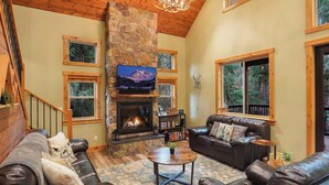 Una sala de estar acogedora con chimenea de piedra, sofás de cuero, televisor y una escalera.