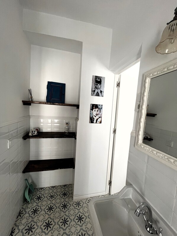 A bathroom with white tiled walls, a patterned floor, a bathtub, a sink, and shelves with toiletries.