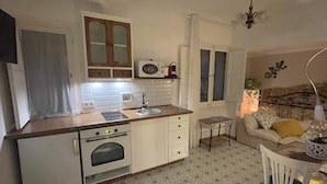 Cocina compacta con armarios blancos, microondas y horno. Hay una pequeña mesa con una lámpara y una silla. El piso presenta un diseño de baldosas con motivos.