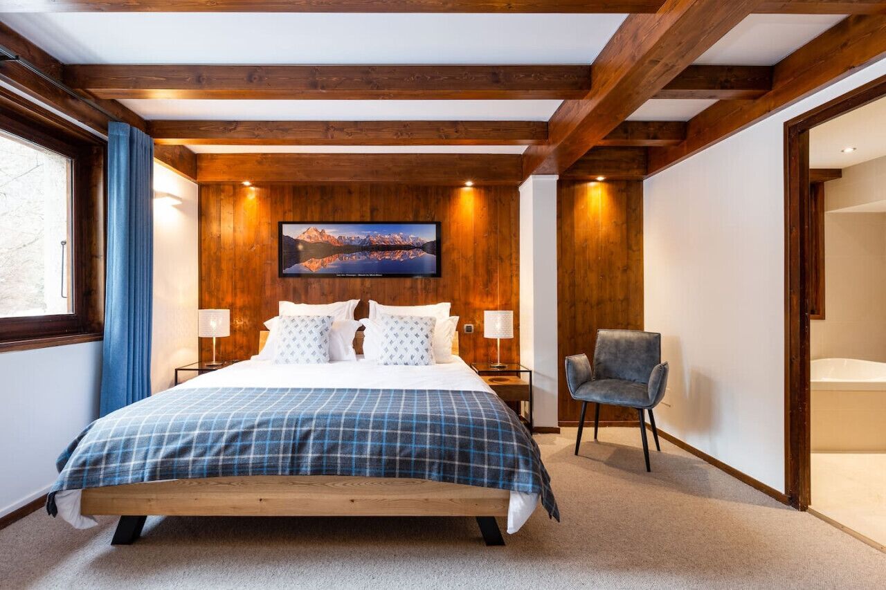Une chambre avec un plafond en bois, un lit recouvert d’une couverture à carreaux, une chaise et un téléviseur fixé au mur qui diffuse une image de paysage montagneux.