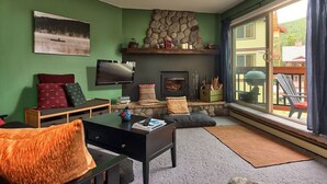 A cozy living room with a stone fireplace, a television, and a large window with a view of the outdoors. - Apex Mountain ~ Beaconsfield Getaway (Apex Mountain Resort)