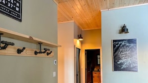 Un pasillo con techo de madera, un banco, un estante con herramientas y una puerta que da a una zona de cocina, además de un cartel con una frase inspiradora.