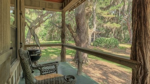 A covered porch with wicker furniture, a grill, and a view of a wooded area. - 3BR Chachalaca Retreat on Sapelo w/ firepit, screened-in porch, & beach supplies (Sapelo Island)