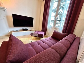 A living room with a large flat-screen TV mounted on the wall, a sofa, a small side table, and a window with curtains. - Flat Duplex Confort T2 Paris Porte De Versailles (Vanves)