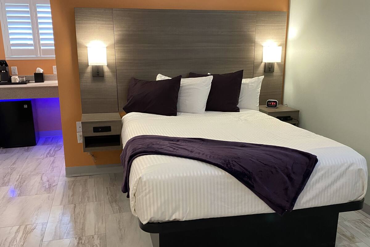 A modern hotel room with a large bed, two bedside lamps, a nightstand with a clock, and a built-in desk with a coffee maker.