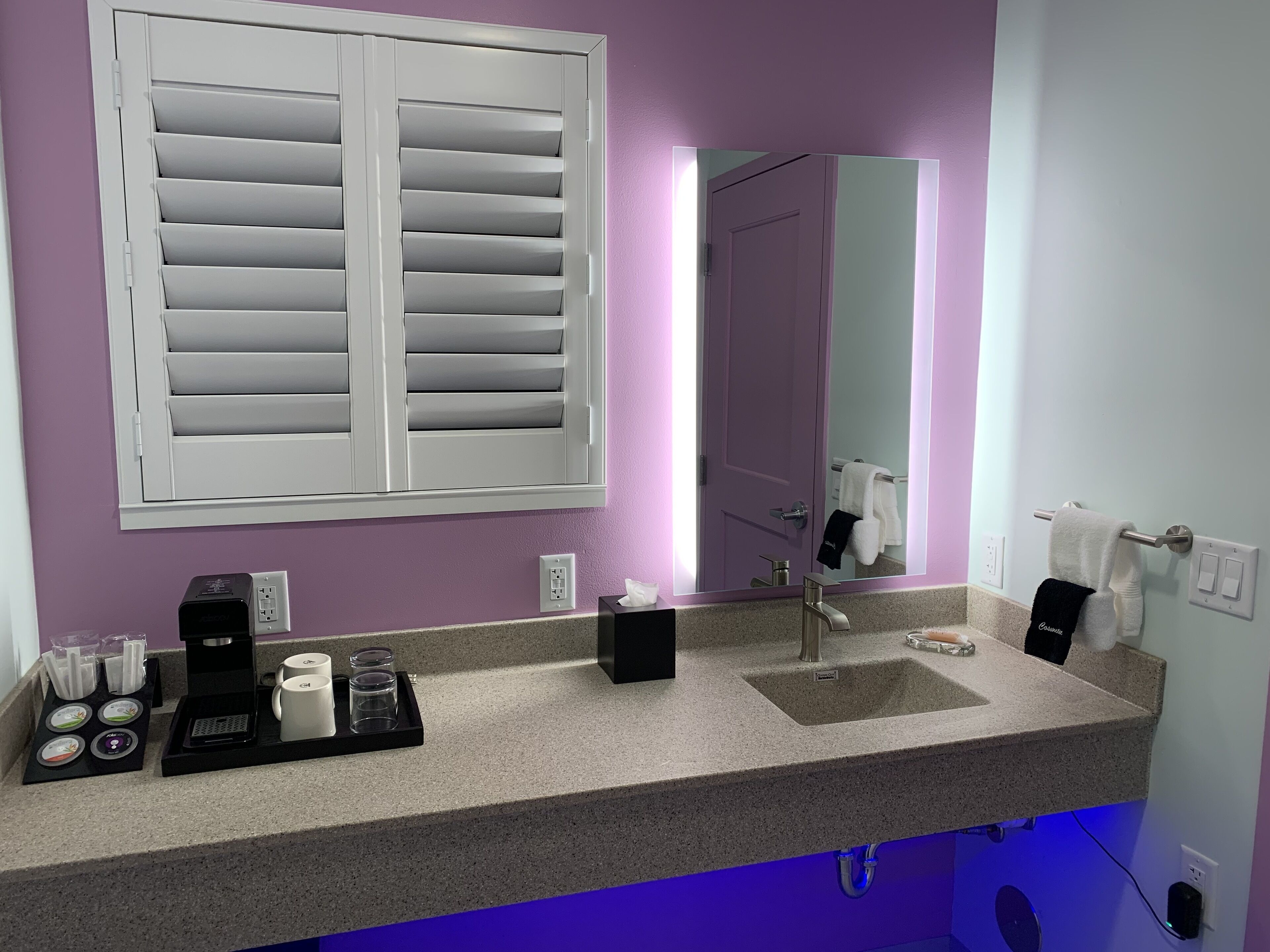 A bathroom with a purple wall, a large mirror, a sink with a faucet, a towel rack, and a window with white blinds.