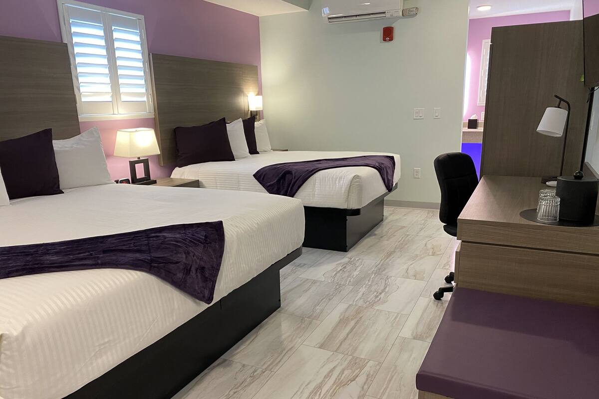 A hotel room with two beds, a desk, a chair, a lamp, and a window with blinds.