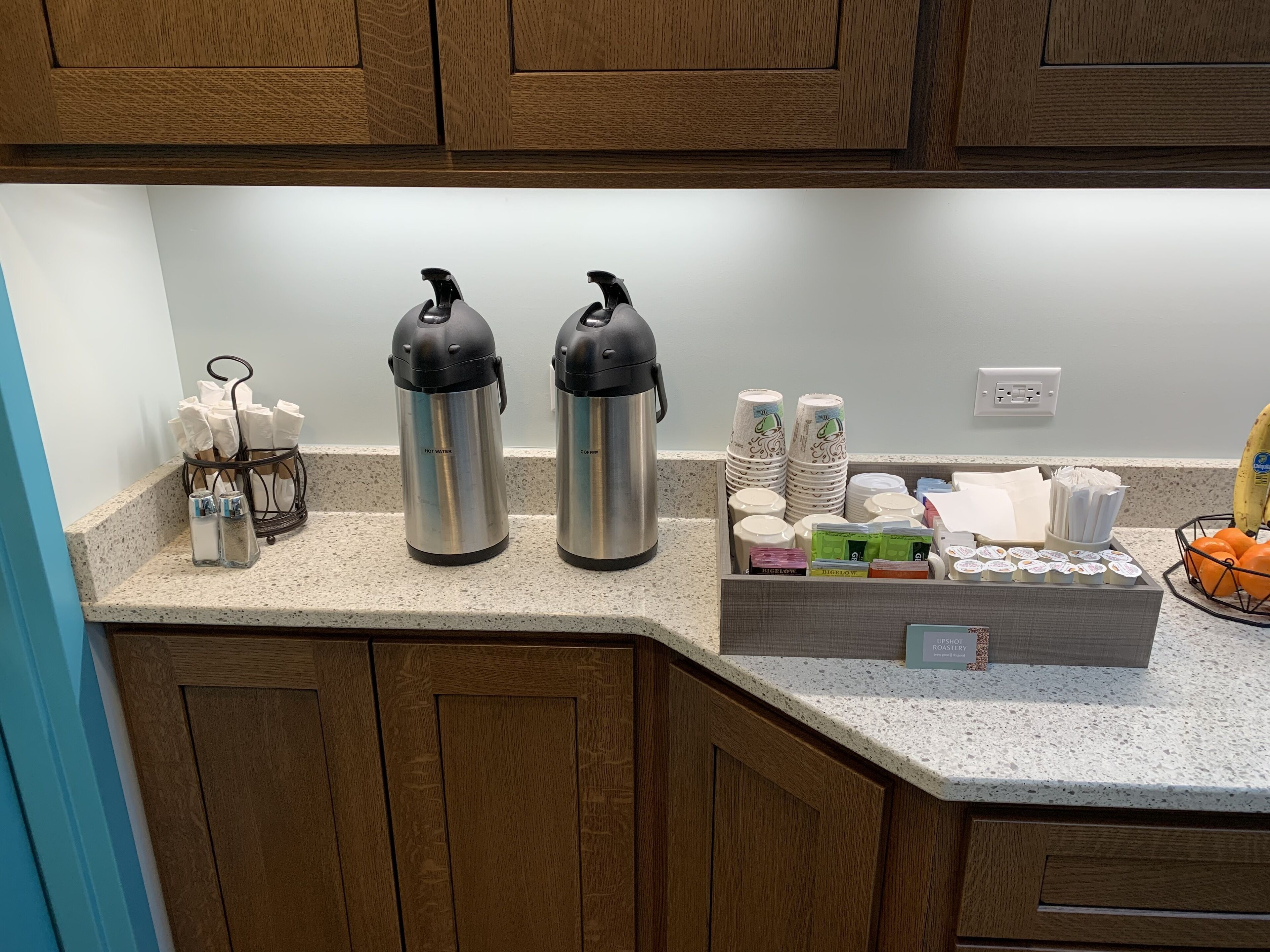 A kitchen counter with stainless steel hot water and coffee dispensers, stacked paper cups, and a box of coffee pods.