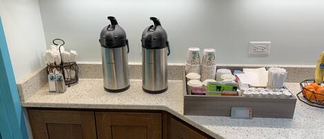 Eine Küchenarbeitsplatte mit Edelstahl-Warmwasser- und Kaffeeautomaten, gestapelten Papierbechern und einer Kapselbox für Kaffee.