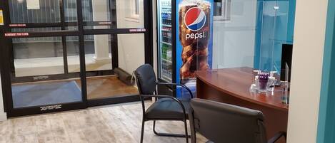 A room with a wooden floor, a desk with two chairs, a glass door with emergency instructions, and a Pepsi vending machine.