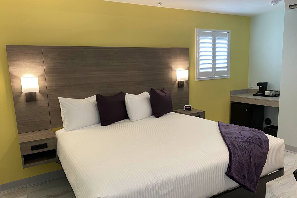 A neatly made bed with white linens and a purple throw blanket, a nightstand with a lamp, and a small black coffee maker on a counter.